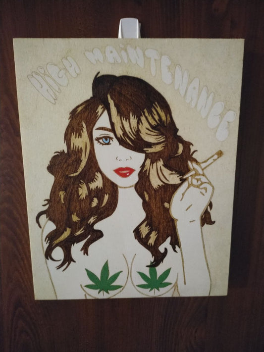 A handburned wooden artwork featuring a stylized woman with wavy hair and red lips, holding a cigarette. Above her, the phrase ‘HIGH MAINTENANCE’ is inscribed. The piece includes two cannabis leaves strategically placed and is mounted on a dark brown wall.