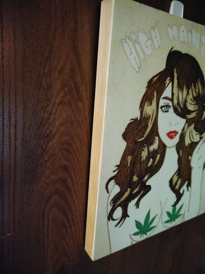 A handburned wooden artwork featuring a stylized woman with wavy hair and red lips, holding a cigarette. Above her, the phrase ‘HIGH MAINTENANCE’ is inscribed. The piece includes two cannabis leaves strategically placed and is mounted on a dark brown wall.